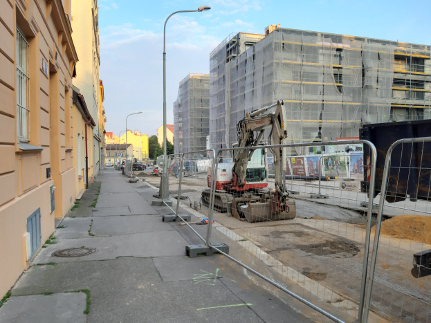 Eine Baustelle mit einem große Bau im Hintergrund, umgeben von Fahrzeugen, Masten, Lichtern, Baümern und einem Zaun.