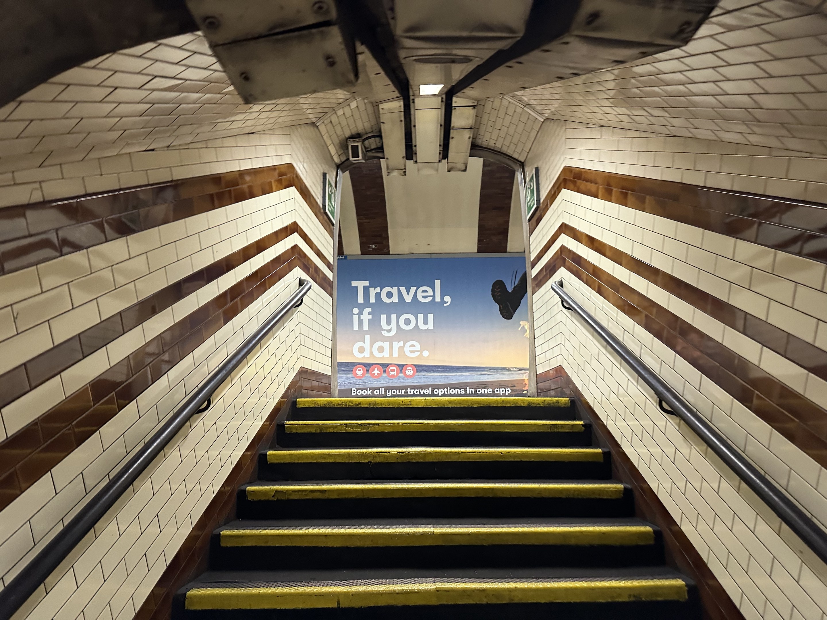 Eine Treppe in einer U-Bahn-Station mit einem 'Reise, wenn du wagst'-Schild oben, flankiert von Geländern und StationWsänden im Hintergrund.