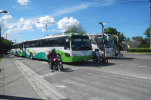 Grüner und weißer Shuttlebus am Straßenrand geparkt mit Motorradfahrern davor, Fußweg mit Gras und Pflanzen links, und Gebäude, Bäume, Laternenmasten und einen klaren blauen Himmel im Hintergrund.