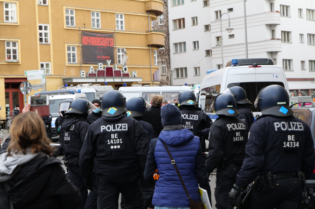 Eine Gruppe von Polizisten vor einer Menge bei einer Demonstration in Berlin, mit Fahrzeugen, Gebäuden und einer Person mit einer Kamera im Hintergrund.
