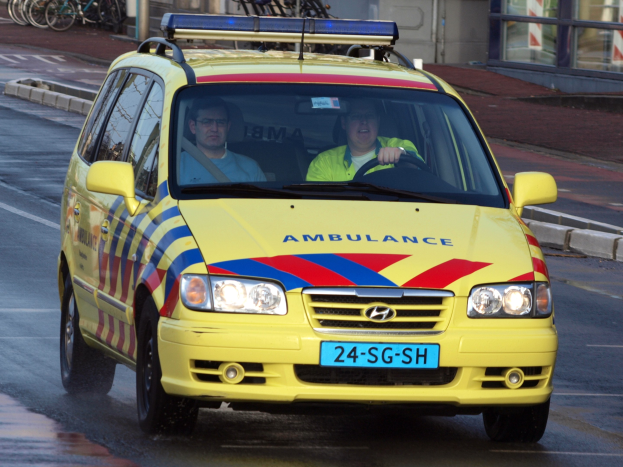 Gelber Krankenwagen fährt auf einer Straße mit zwei Personen darin, Fahrräder auf dem Gehweg zur Linken und ein Gebäude mit Glasfenstern im Hintergrund.