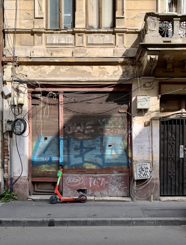 Ein roter Roller neben einem Gebäude mit Fenstern, Rollläden und einem Tor auf einer Straße mit sichtbaren Kabeln und Gegenständen, darunter Text am unteren Rand.