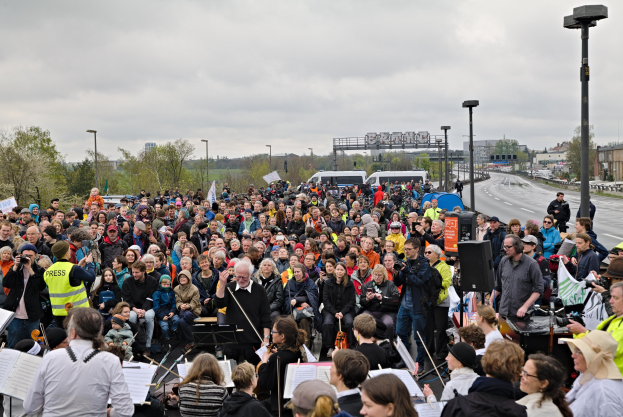 Eine große Gruppe von Menschen marschiert vor einer Menge, einige halten Musikinstrumente, Stände und andere Gegenstände, mit Fahrzeugen, Laternenmasten, Bäumen, Gebäuden und einem bewölkten Himmel im Hintergrund.
