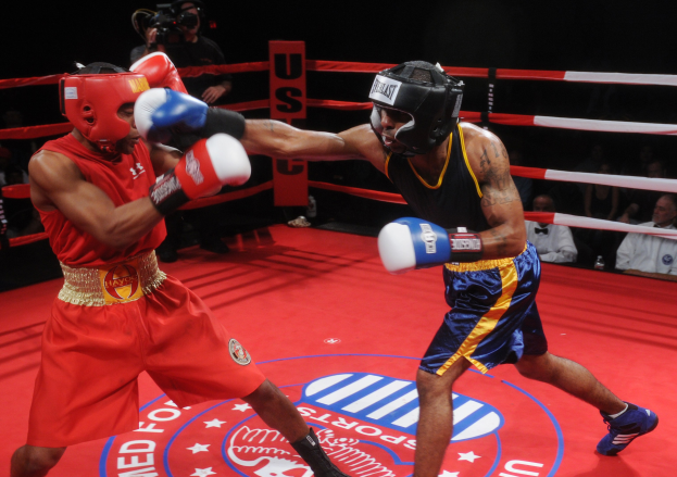 Zwei Boxer kämpfen im Ring, während ein Mann im Hintergrund filmt, mit einer kleinen Zuschauerschaft in gedämpfter Beleuchtung.