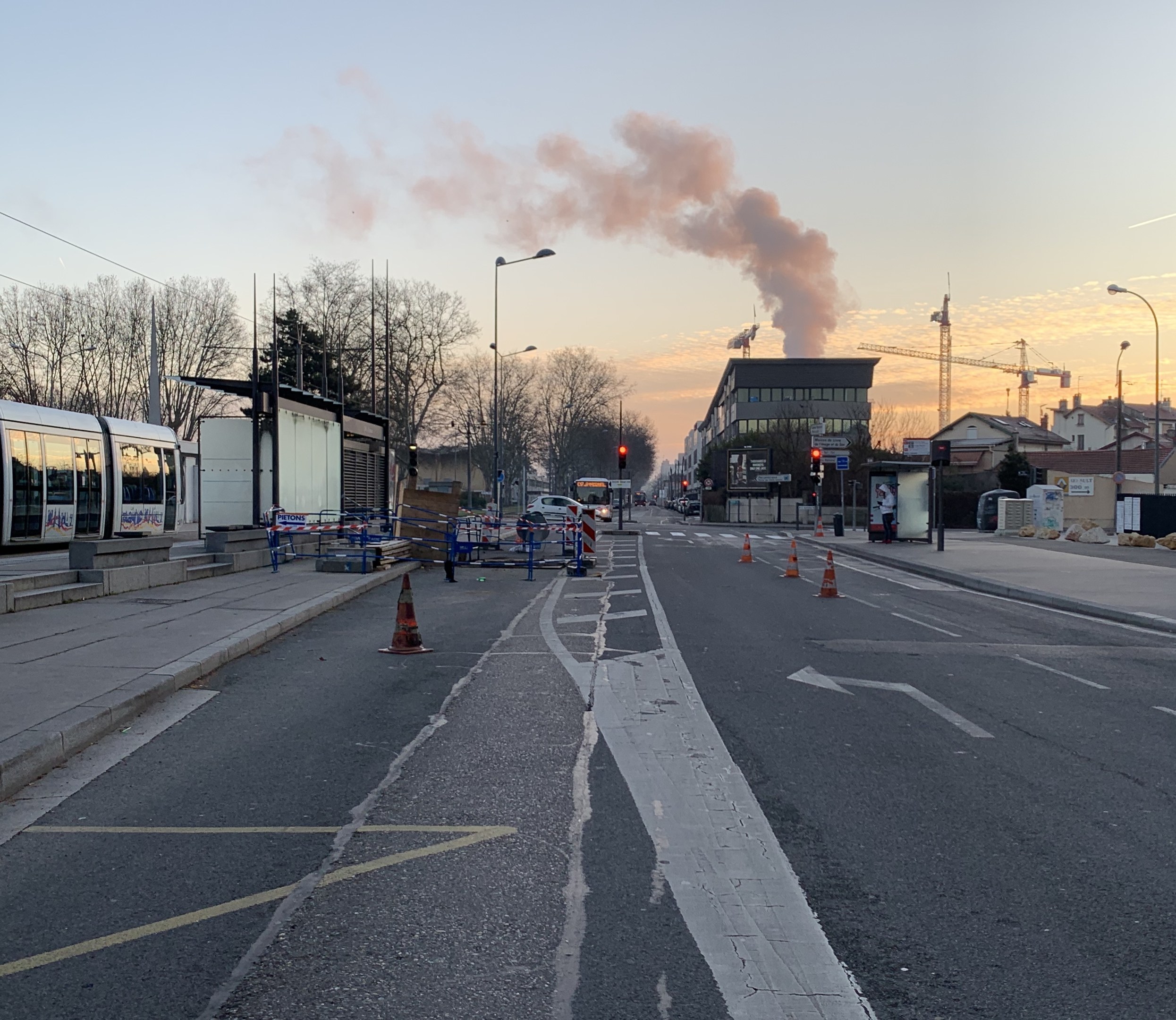 Eine städtische Straße mit Absperrbaken, einem Bus, Laternen, Bäumen, Gebäuden, Kränen und Rauch, der aus einem fernen Gebäude aufsteigt.