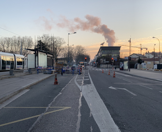 Eine städtische Straße mit Absperrbaken, einem Bus, Laternen, Bäumen, Gebäuden, Kränen und Rauch, der aus einem fernen Gebäude aufsteigt.
