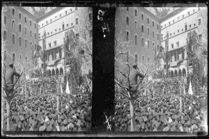 Ein Schwarz-Weiß-Foto einer großen Menschenmenge vor einem Gebäude mit Fenstern, Bäumen und Fahnen im Hintergrund, die scheinbar eine Protestveranstaltung mit einigen Schildern und Transparenten abhalten.