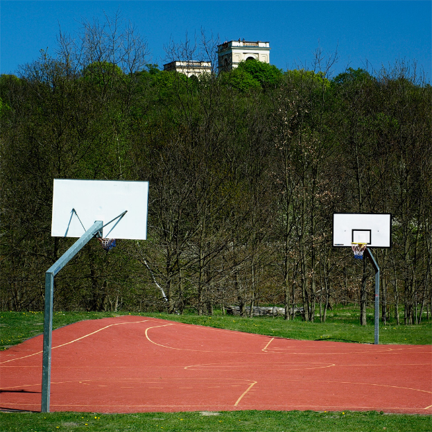 Ein Spielplatz mit Gras, zwei Basketballkörben, Bäumen, einem Gebäude im Hintergrund und einem klaren Himmel.