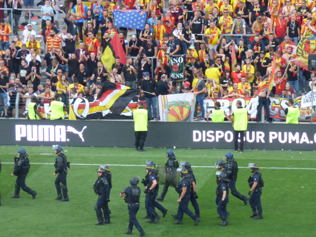 Eine Gruppe von Polizisten in Helmen und mit Waffen in der Hand läuft über ein Fußballfeld, im Hintergrund halten Menschen Fahnen und Banner in der Nähe von Geländern.