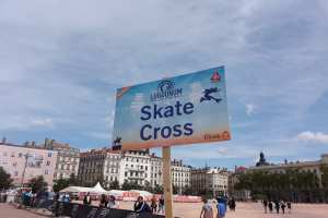 Großes Werbeplakat auf einem Stadtplatz umgeben von Menschen, mit Schildern, sitzenden und stehenden Gruppen, einem Zaun, Zelten, Straßenmöbeln, Bäumen, Gebäuden und einem bewölkten Himmel.