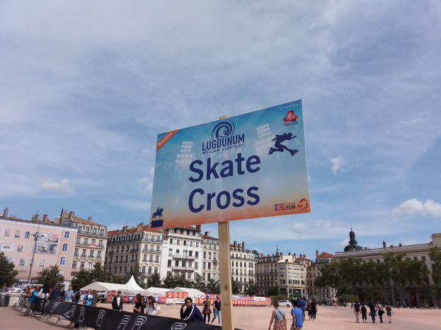 Großes Werbeplakat auf einem Stadtplatz umgeben von Menschen, mit Schildern, sitzenden und stehenden Gruppen, einem Zaun, Zelten, Straßenmöbeln, Bäumen, Gebäuden und einem bewölkten Himmel.