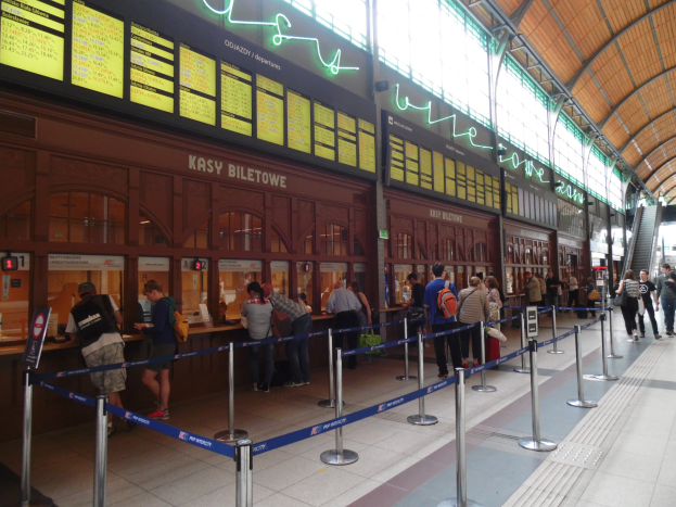 Menschen in einer Warteschlange an einem Bahnhofsschalter mit Taschen, Stangen mit Bändern, Informationsschildern, Deckenleuchten und Treppen mit Gelöndern.