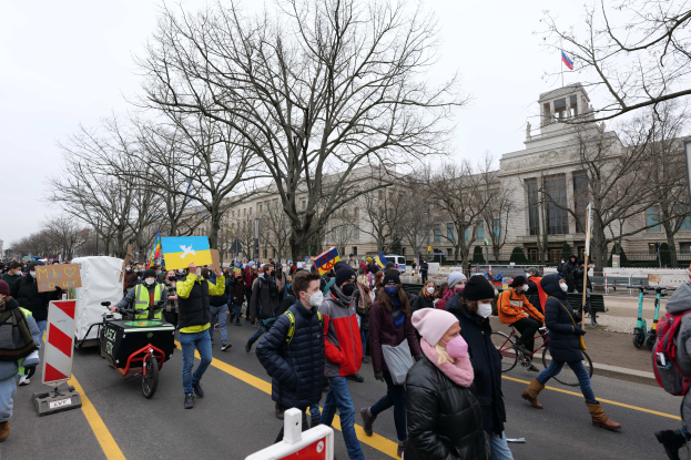 Eine große Protestmarsch mit Menschen, die gehen, Schilder halten und Fahrräder fahren, auf einer Straße in Washington, D.C., mit Bäumen, Schildern und einem klaren blauen Himmel im Hintergrund.