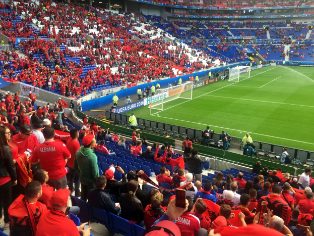 Eine große Menschenmenge in einem Stadion schaut einem Fußballspiel zu, mit Menschen auf Stühlen und Stehplätzen, Bannern, einem Torpfosten und Deckenlampen.