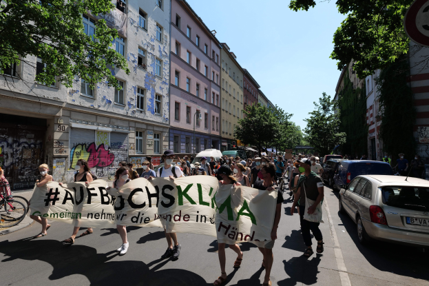 Eine Gruppe von Menschen marschiert mit einem Transparent die Straße entlang, mit parkenden Autos auf der rechten Seite, einem Fahrrad auf der linken Seite und Gebäuden, Bäumen und einem Schild im Hintergrund unter einem sichtbaren Himmel.