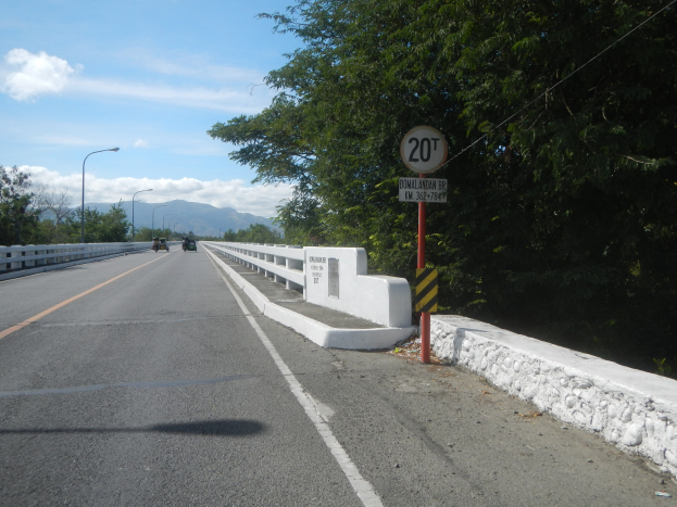 Eine Fahrspur mit Fahrzeugen, ein Tempolimit-Schild, Geländer, Laternen, Bäume und ein bewölkter Himmel im Hintergrund.