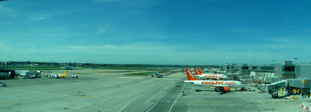 Eine Flughafen-Szene mit Flugzeugen, Gebäuden, Masten, Fahrzeugen, Bäumen und einem Himmel darüber sowie einer Straße darunter und einem Zaun im Vordergrund.