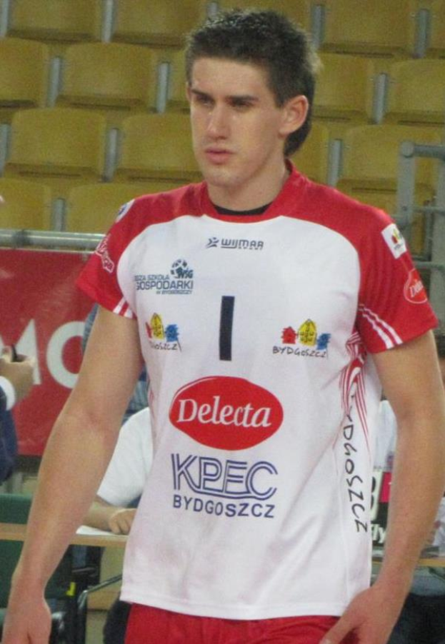 Ein Mann in einem roten und weißen Handballtrikot steht auf einem Platz, umgeben von Zuschauern, mit einem roten Banner im Hintergrund, auf dem "Krasnoyarsk vs Bydgoszcz" steht, und sieht entschlossen aus.