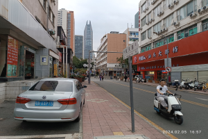 Ein Mann fährt mit einem Roller eine Straße in der Stadt entlang, gesäumt von hohen Gebäuden, mit Menschen auf den Gehwegen und anderen Fahrzeugen auf der Straße, unter einem klaren blauen Himmel mit Bäumen im Hintergrund.
