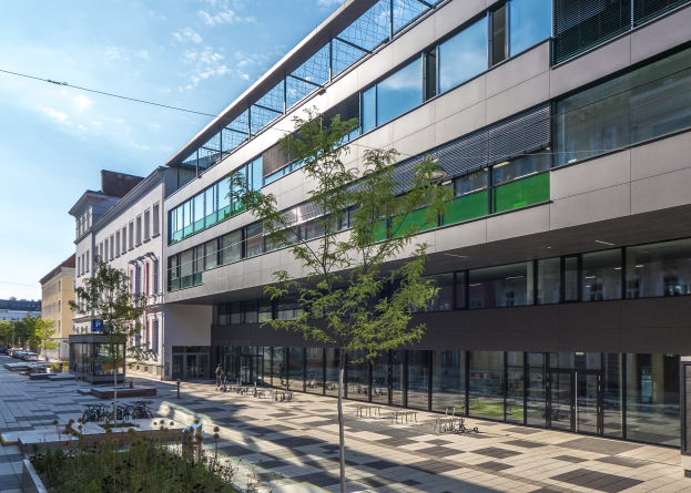 Großes modernes Gebäude mit vielen Fenstern, umgeben von Bäumen und Grünflächen, mit Fahrzeugen und Menschen in der Nähe, unter einem bewölkten Himmel, als das neue deutsche Institut für Technologie-Hauptquartier in Berlin identifiziert.