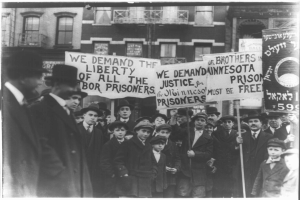 Schwarzes und weißes Foto einer Gruppe von Menschen in einer Reihe mit Schildern, auf denen "Wir fordern die Freiheit aller Gerechtigkeit für die Gefangenen" steht, vor einem Gebäude mit Fenstern und einem Geländer.