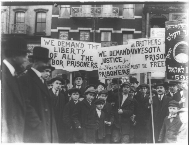 Schwarzes und weißes Foto einer Gruppe von Menschen in einer Reihe mit Schildern, auf denen "Wir fordern die Freiheit aller Gerechtigkeit für die Gefangenen" steht, vor einem Gebäude mit Fenstern und einem Geländer.