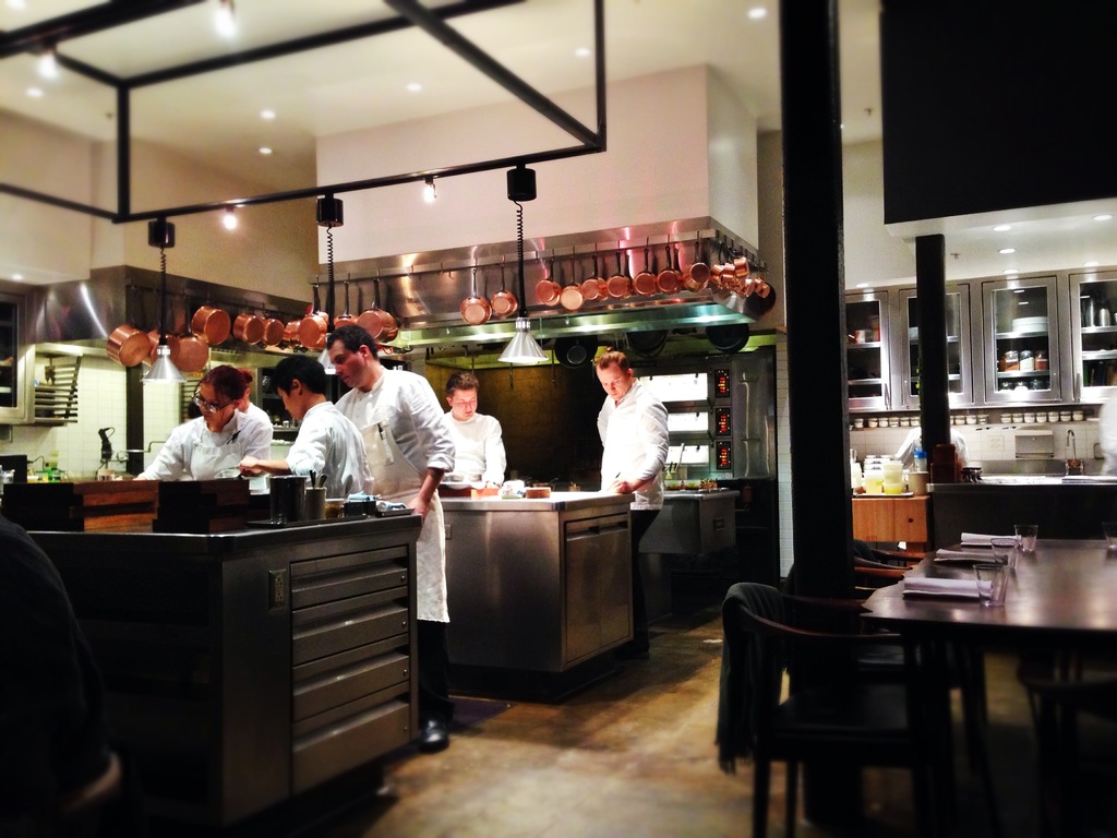 Ein Holztisch in einer Küche mit einem Glas und Papieren darauf, umgeben von fünf arbeitenden Personen.