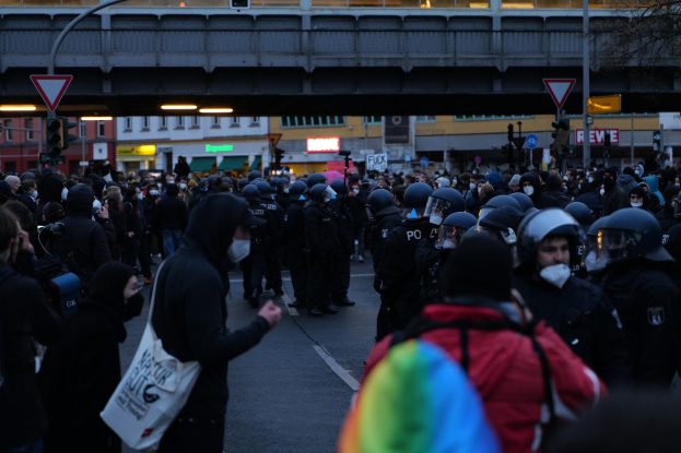 Eine große Gruppe von Menschen steht einer Reihe von Polizisten gegenüber, einige tragen Helme und tragen Taschen oder Kameras, mit Gebäuden, Schildern, Pfosten, einer Brücke und einem Baum im Hintergrund.