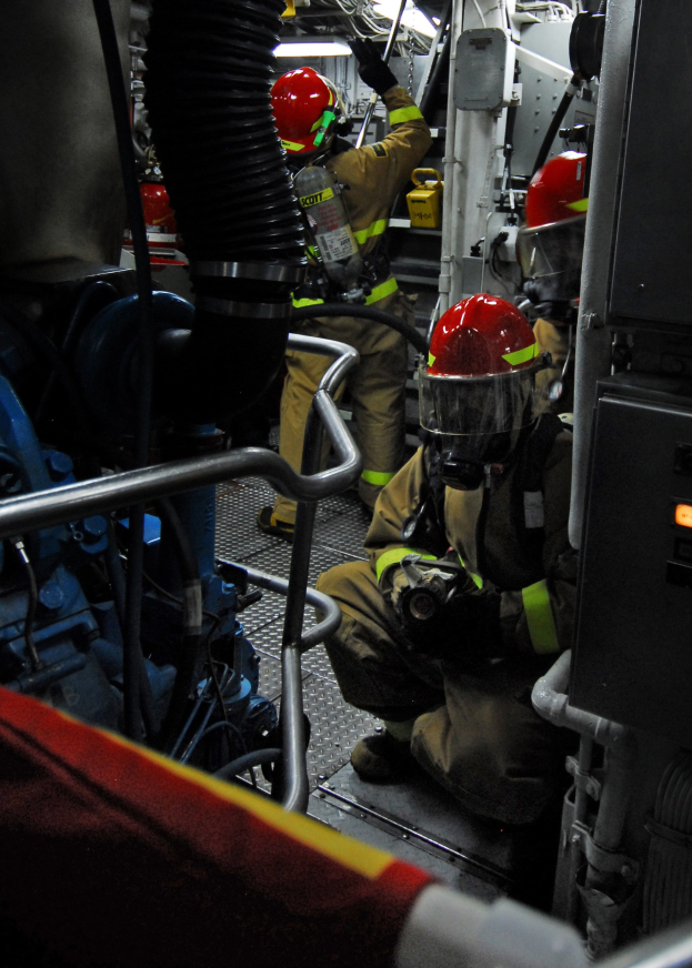 Feuerwehrleute in Helmen und Schutzausrüstung arbeiten an einem Feuerwehrauto mit Rohren und Gegenständen im Hintergrund, unter einem Licht an der Spitze.