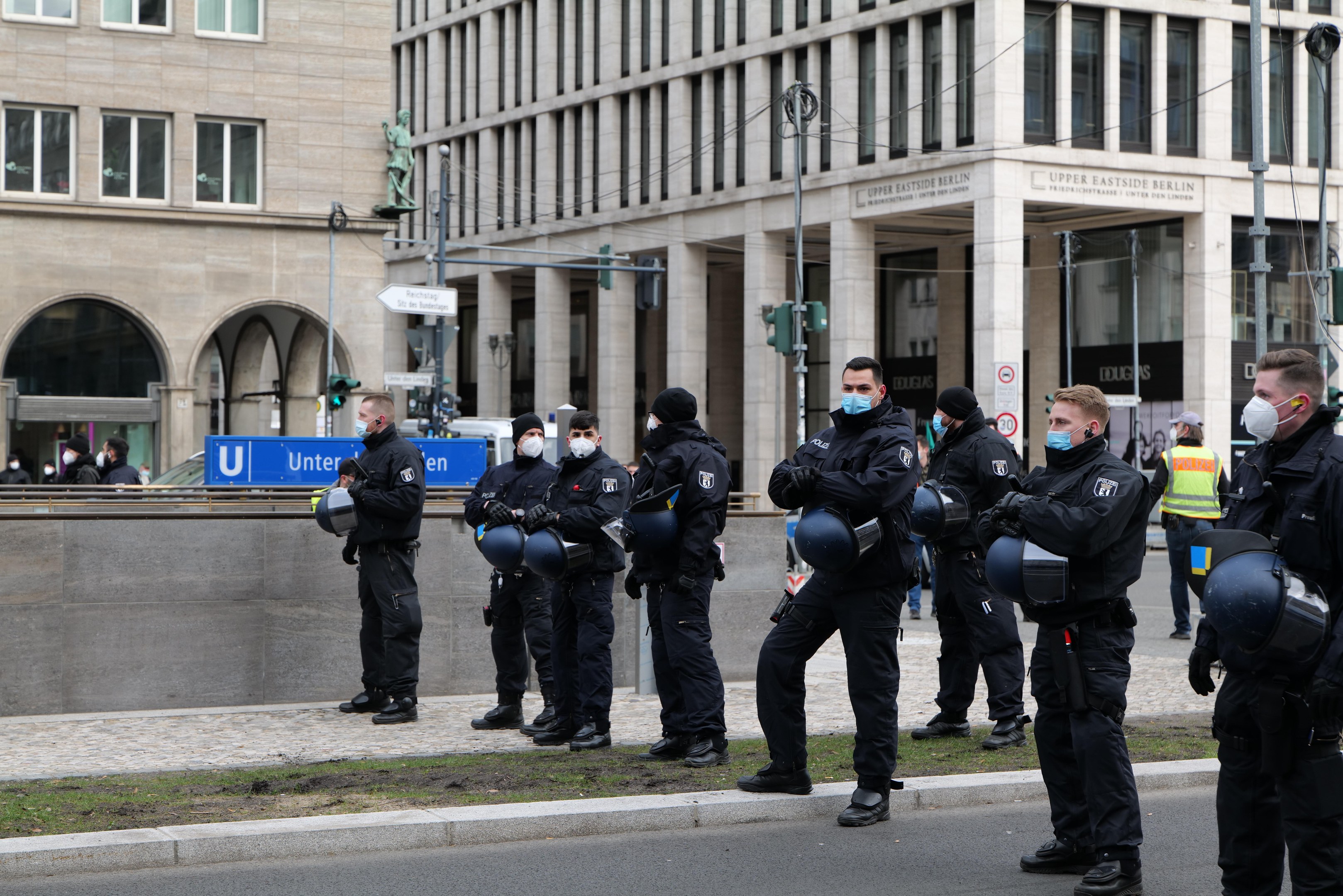 Eine Gruppe von Polizisten in schwarzen Uniformen und Masken steht vor einem Berliner Gebäude mit Glasfenstern, Säulen und Gras, einige halten Helme, mit Laternen, Verkehrszeichen, Schildern mit Text, einer Statue und anderen Menschen im Hintergrund.