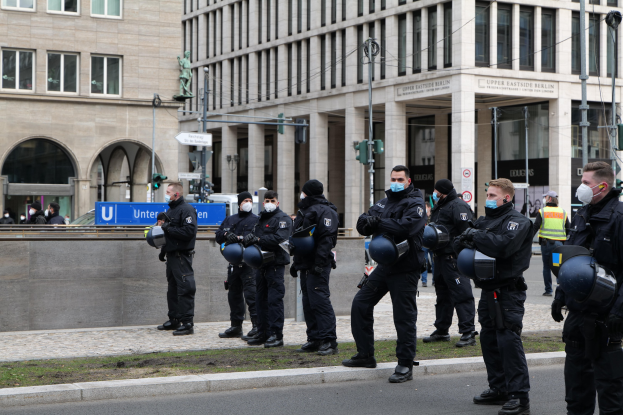Eine Gruppe von Polizisten in schwarzen Uniformen und Masken steht vor einem Berliner Gebäude mit Glasfenstern, Säulen und Gras, einige halten Helme, mit Laternen, Verkehrszeichen, Schildern mit Text, einer Statue und anderen Menschen im Hintergrund.