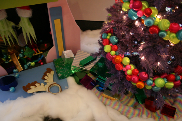 Innen: Ein geschmückter Weihnachtsbaum mit Lichterkette und Schmuck steht rechts, Geschenkboxen und Dekorationen in der Mitte, eine Hütte mit Dekorationen oben links und Baumwolle und Wolle unten.