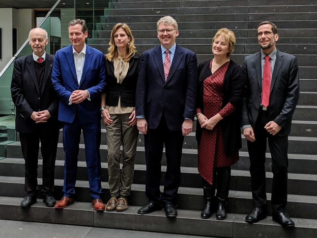 Mitglieder des Europäischen Parlaments stehen zusammen auf Treppenstufen, lachend, mit einer Glastür und einer Treppe mit Geländer im Hintergrund.