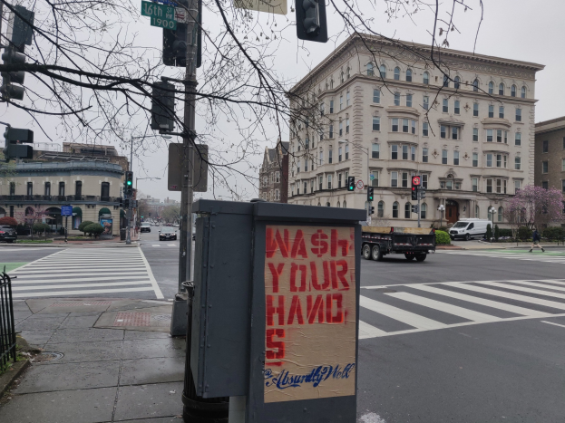 Eine belebte Stadtstraße mit fahrenden Fahrzeugen, einem Fußgänger auf dem Gehweg, einem Müllcontainer mit der Aufschrift "Hände Waschen", Verkehrsampeln, Straßenschilder, einem Metallzaun, Grünpflanzen, Gebäuden und einem blauen Himmel.