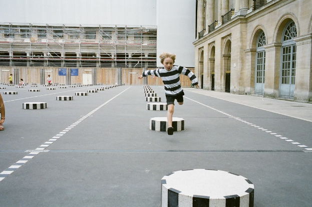 Ein Spielplatz mit vier Kindern, eines läuft auf einem schwarz-weißen Betonstufenbereich, umgeben von einem Schulgebäude.