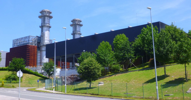 Ein großes Gebäude mit zahlreichen Fenstern, das durch eine Beschilderung als Kraftwerk identifiziert wird, umgeben von Straßeninfrastruktur, Fahrzeugen, Grünflächen und einem bewölkten Himmel.