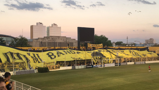 Ein Fußballfeld mit einer Gruppe von Menschen darauf, umgeben von einem Metallzaun, einem Torpfosten, einer Banner mit der Aufschrift "Boca Juniors vs Real Sociedad", einigen Pflanzen, einem Anzeigebildschirm, einigen Pfosten, der Rinde eines Baumes, einigen Bäumen, einigen Gebäuden und einem bewölkten Himmel.