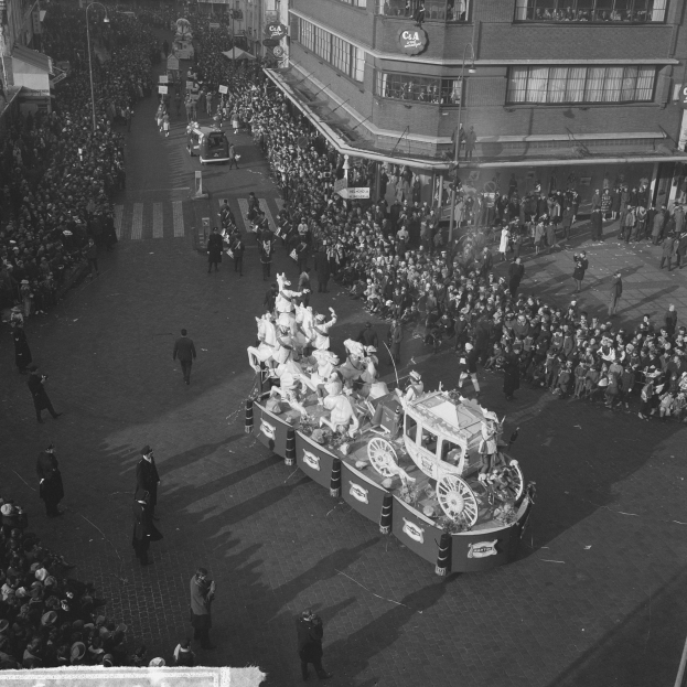 Eine große Menschenmenge umringt einen Schmuckwagen bei einem Umzug, der mit bunten Luftballons und Bändern geschmückt ist, mit Gebäuden und Laternen im Hintergrund, in Schwarz-Weiß.