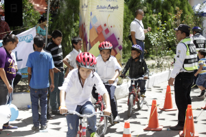 Gruppe von Kindern, die Fahrräder auf einer Straße mit Verkehrskegeln fahren, einige tragen Helme, andere stehen in der Nähe, mit einem Banner, Bäumen und Gebäuden im Hintergrund.