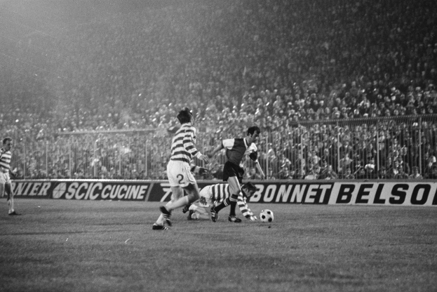 Eine Gruppe von Männern, die Fußball auf einem Feld spielen, mit Zuschauern auf den Tribünen und einem Banner mit der Aufschrift "Celtic v West Bromwich Albion - FA Cup Third Round" in Schwarz-Weiß.