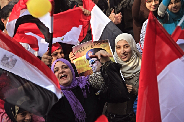 Eine Gruppe ägyptischer Demonstranten auf dem Tahrir-Platz in Kairo, die Fahnen schwenken und Parolen rufen, während einige Schals tragen und einer ein Schild hält, vor einem Hintergrund mit einem Ballon.