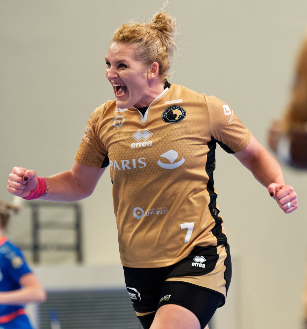 Eine Frau in braunem T-Shirt und schwarzen Shorts rennt auf einem Futsalplatz, feiert mit triumphaler Miene, während zwei andere Frauen im Hintergrund unscharf sind.