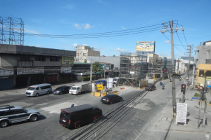 Eine belebte Stadtstraße mit zahlreichen Fahrzeugen, hohen Gebäuden, Ampeln, Strommasten mit Drähten, Texttafeln, Bäumen und einem bewölkten Himmel.