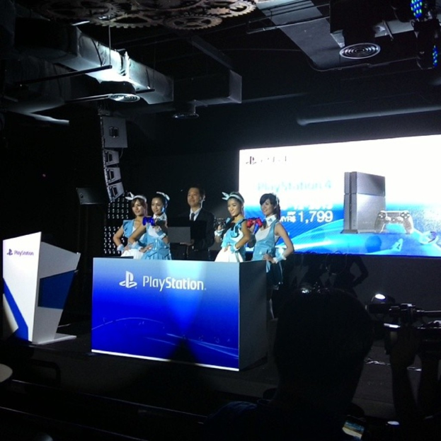 Menschen auf einer Bühne in der Nähe eines Podiums mit "Playstation" darauf, mit einem Projektionsschirm an der Wand dahinter.