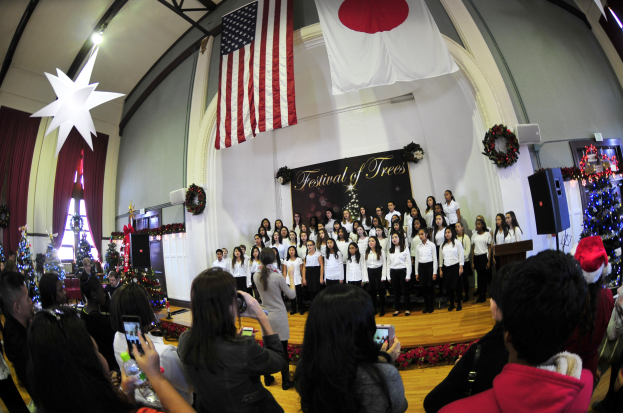 Eine Gruppe von Menschen auf einer Bühne vor einem Publikum, mit Weihnachtsdekoration, einem Podium, Lautsprechern, einer Texttafel, Vorhängen, Flaggen und Deckenleuchten.