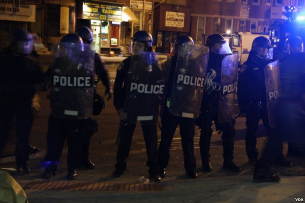 Polizeibeamte in Helmen und Schilden stehen in einer Straße bei Nacht, mit Gebäuden, Fahrzeugen und Laternen im Hintergrund.