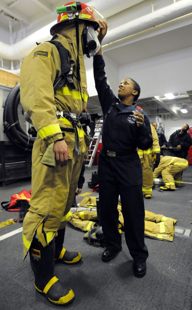 Zwei Feuerwehrleute, eine in schwarzer Uniform und Helm und ein anderer in gelb-schwarzer Ausrüstung, stehen in der Nähe einer Leiter und Rohre mit anderen im Hintergrund.