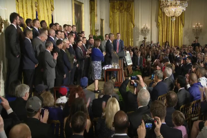 Eine Gruppe von Menschen mit Kameras steht vor einer Menge, einem Podium mit Mikrofon und Texttafel, einer Topfpflanze, Kerzen, Rahmen, Vorhängen und einem Kronleuchter und fotografiert den Präsidenten und Vizepräsidenten der Vereinigten Staaten.