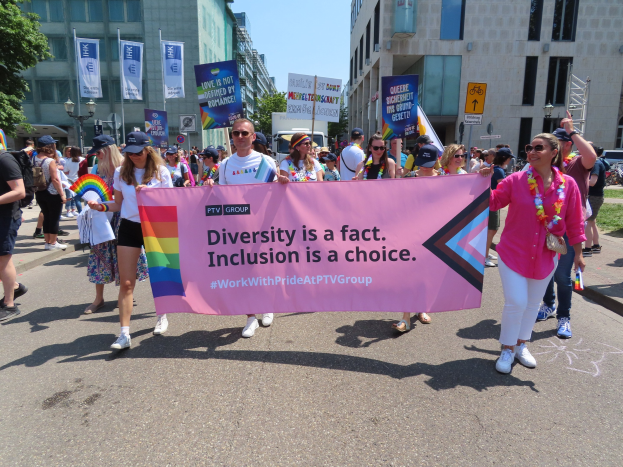 Eine Gruppe von Menschen marschiert auf einer Straße, hält ein Banner mit der Aufschrift "Diversität ist eine Tatsache, Inklusion ist eine Wahl" und trägt teilweise Mützen und Schutzbrillen, während andere zusätzliche Banner halten, vor einem Hintergrund aus Gebäuden, Bäumen und einem klaren blauen Himmel.