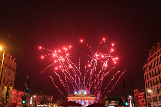 Eine Straße in Berlin an Silvester, voller Gebäude, Bäume, Laternenmäste, Verkehrszeichen, Zelte und Menschen, mit einem Feuerwerk am Himmel im Hintergrund.